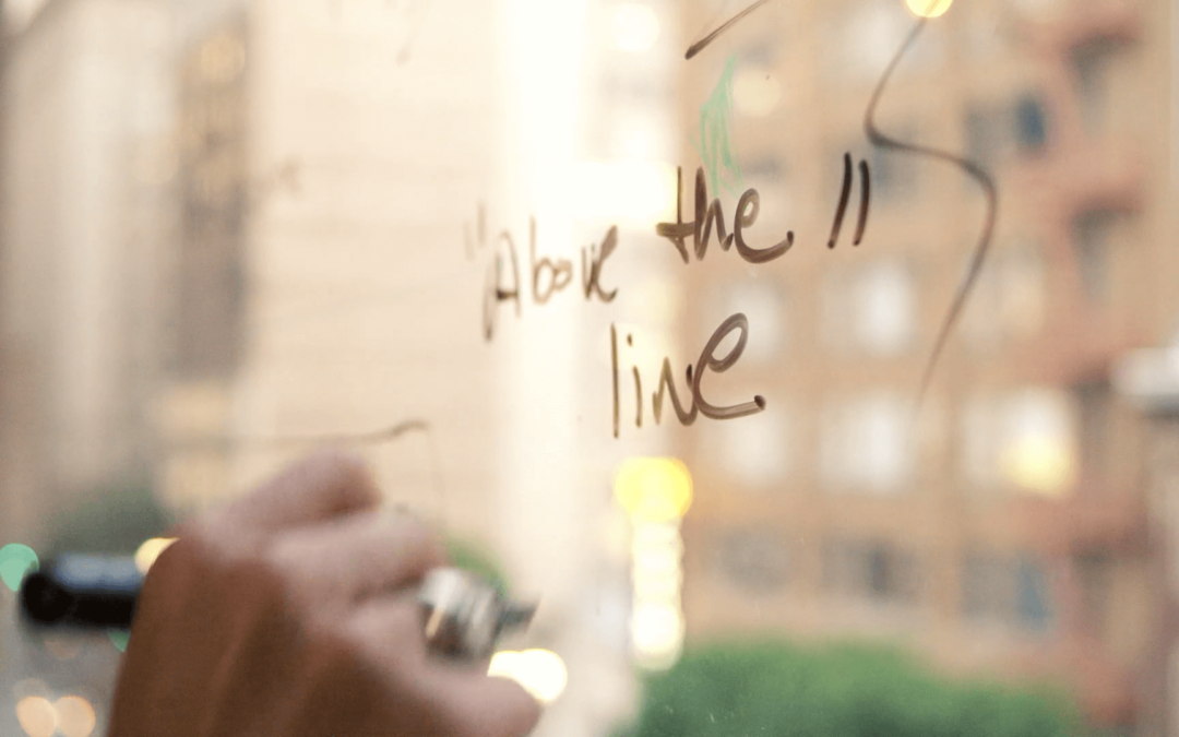 person writing with marker on a window, reads "above the line"